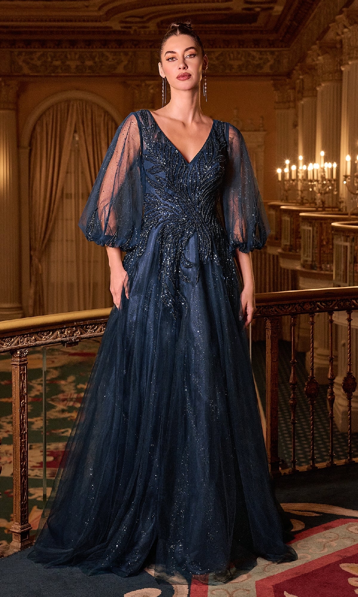 A woman poses indoors in the Ladivine CD0101 Balloon-Sleeve Blue Formal Gown, a shimmering navy blue, floor-length dress with glitter accents and translucent balloon sleeves, elegantly showcased beneath ornate chandeliers.